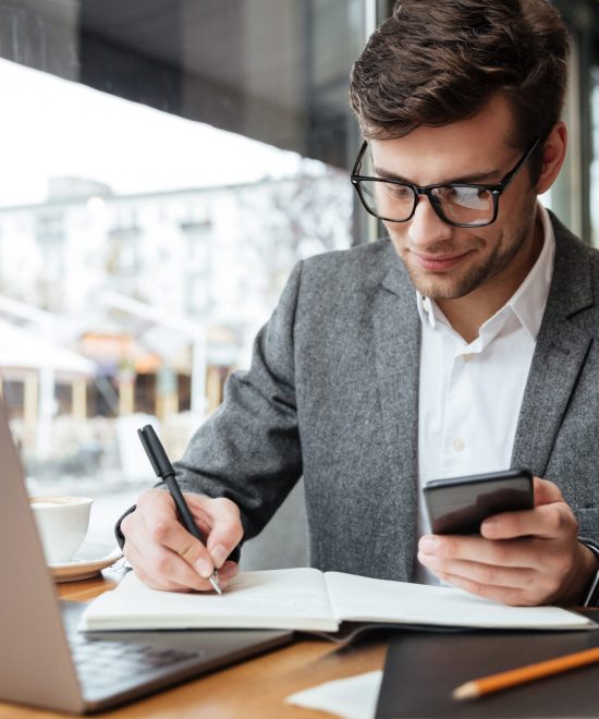 Smiling business man in eyeglasses sitting by the table in cafe with laptop computer while using smartphone and writing something